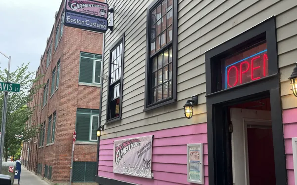 Gray and pink building with lamp lights and a neon "OPEN" sign above the door. Two signs protrude from the building, saying "The Garment District" and "Boston Costume" 