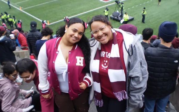 Two best friends smiling at the camera in the stands at Harvard vs Yale game.