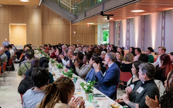 Many students, faculty, and staff sit at several long tables, eating and talking