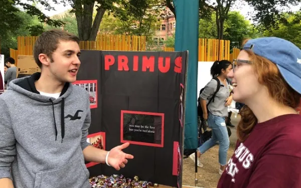 two people talking outside next to a poster that says Primus